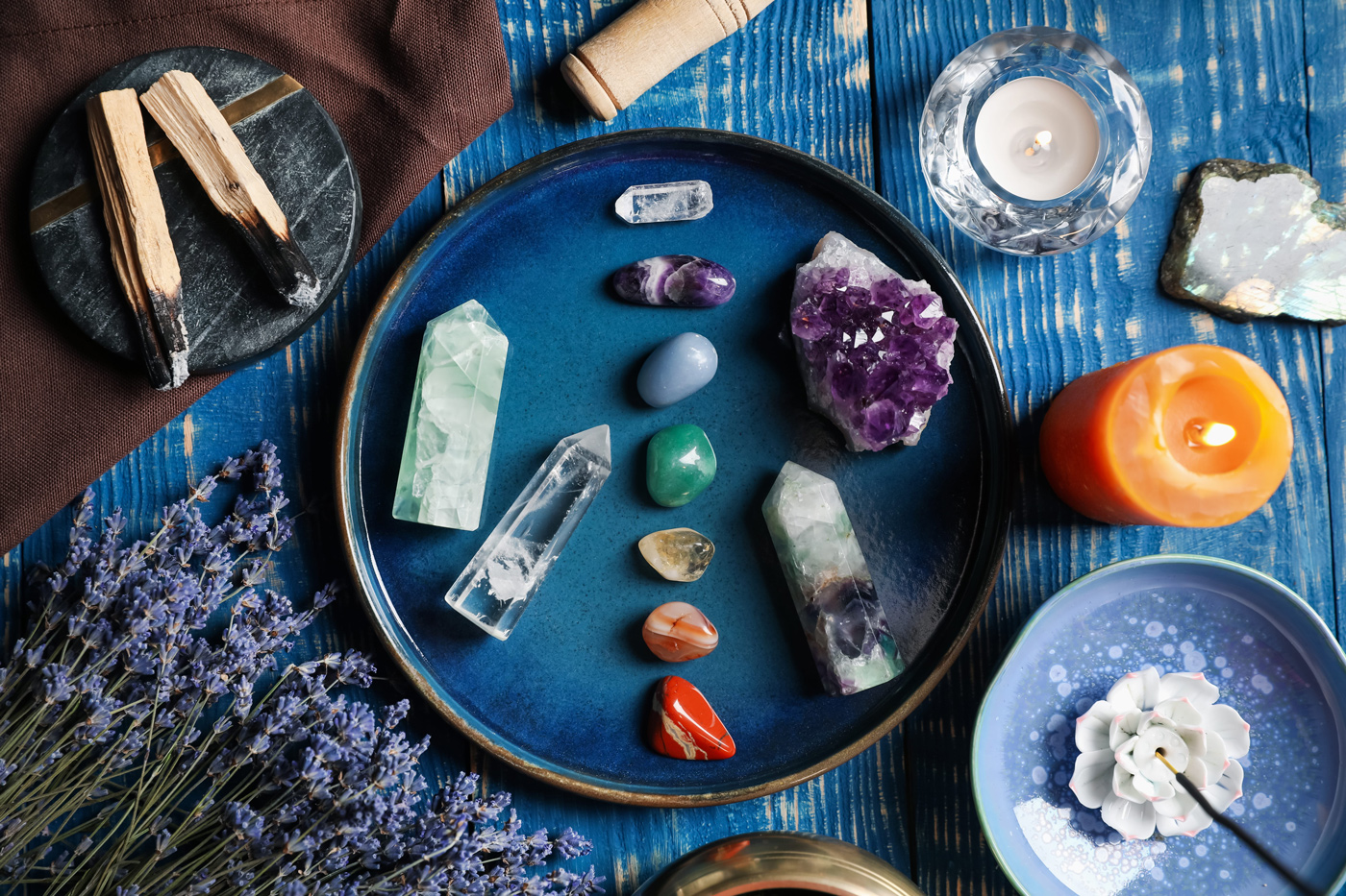 A clear quartz crystal, amethyst geode, several colored gemstones, dried lavender, and multiple candles burn on a table.